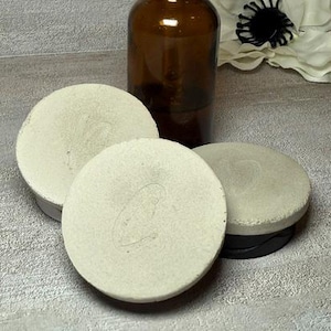 May include: A brown glass dropper bottle with a black top sits next to three round, light-colored objects. A white flower with a black center is in the background. The objects appear to be cosmetic or skincare related.