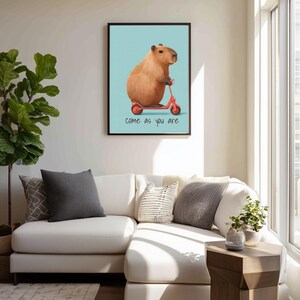 May include: A framed art print featuring a capybara riding a red scooter against a light blue background. The print includes the phrase "come as you are" in black text. The artwork is displayed above a white sectional sofa.