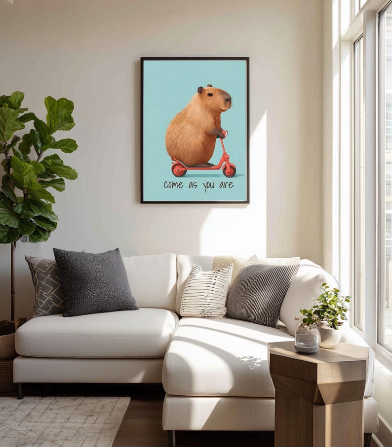 May include: A framed art print featuring a capybara riding a red scooter against a light blue background. The print includes the phrase "come as you are" in black text. The artwork is displayed above a white sectional sofa.