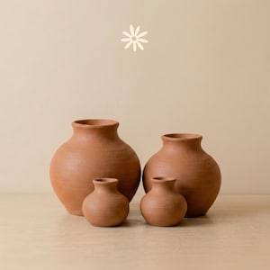 Puede incluir: Una colección de cuatro jarrones de terracota de distintos tamaños, con cuerpo redondeado y cuello estrecho. El conjunto incluye dos jarrones más grandes y anchos y dos más pequeños y compactos, todos con un acabado mate de color tierra.