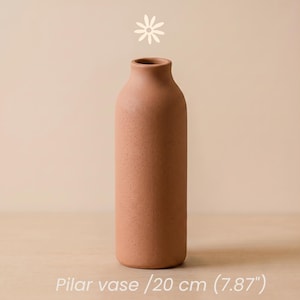 Puede incluir: Un jarrón cilíndrico de color terracota con cuello estrecho y acabado mate texturizado. El jarrón mide 20 cm de altura. Un pequeño icono de flor blanca se sitúa sobre el jarrón, y el texto Pilar vase / 20 cm (7.87") aparece debajo.