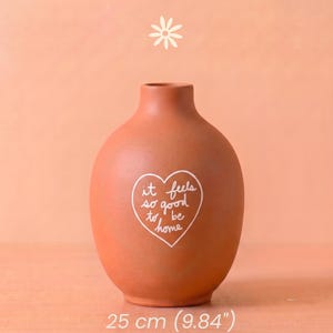 Puede incluir: Un jarrón de cerámica color terracota con cuerpo redondeado y cuello estrecho. En la parte frontal tiene impreso el contorno de un corazón blanco que contiene el texto manuscrito 'it feels so good to be home'. El jarrón mide 25 cm de altura.
