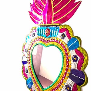 Peut inclure: Miroir en forme de cœur avec un design peint à la main. Le cadre présente un bord festonné avec des couleurs vives, notamment du rose, du bleu, du jaune et du vert. Un motif de flamme stylisé surmonte le miroir.