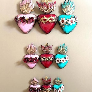 Op de afbeelding: Negen kleurrijke, hartvormige wanddecoraties. Elk hart heeft een vlamontwerp aan de bovenkant en is versierd met bloemen- of kroondetails. De harten zijn geschilderd in roze, rood en turquoise, met gouden accenten.