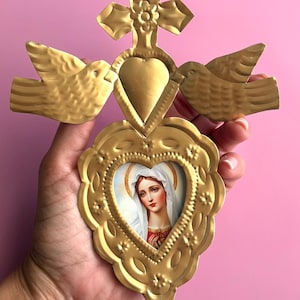 May include: Gold-colored metal religious wall hanging featuring a cross, heart, and two doves. The heart-shaped frame holds a portrait of a woman with a white veil. The piece is held against a pink background.
