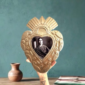 May include: A heart-shaped brass wall hanging featuring an embossed floral pattern and a central heart-shaped frame. The frame contains a black and white portrait of a person with dark hair, wearing a shawl, looking directly at the viewer.