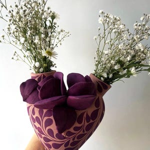 May include: A heart-shaped ceramic vase with a light pink base and a dark purple teardrop pattern. The vase features two openings at the top, each holding white baby's breath flowers, and is adorned with large, dark purple sculpted flower petals.