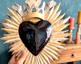 May include: A decorative wall ornament featuring a central dark navy blue heart shape surrounded by a sunburst of alternating gold and silver rays. The top of the heart is adorned with a silver crown-like crest with ornate, leaf-shaped details.
