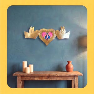 May include: A gold-toned metal wall hanging featuring a central heart-shaped frame with a small image of the Virgin Mary inside, flanked by two stylised birds with textured wings on either side, creating a symmetrical decorative piece.