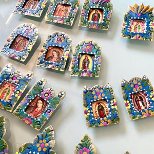 Puede incluir: Una colección de pequeños marcos metálicos rectangulares con motivos florales pintados a mano en azul, verde, rosa y amarillo. Cada marco contiene un retrato religioso en miniatura de una figura con túnica, algunos con remates arqueados.