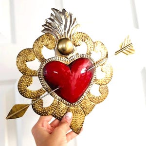 May include: A decorative metal heart with a red center pierced by an arrow. The heart is surrounded by gold filigree and topped with a silver and gold flame design. The heart is held against a white background.