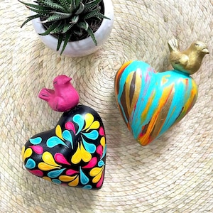 May include: Two heart-shaped sculptures with bird figures on top. One heart is black with colorful teardrop patterns in yellow, pink, and blue. The other heart features a vibrant drip-paint design in turquoise, orange, and gold with a gold bird.