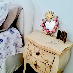 Peut inclure: Une table de chevet crème à deux tiroirs, ornée de motifs floraux dorés. Un miroir en forme de cœur avec des accents de cœurs roses et un motif de flamme dorée est posé sur la table de chevet, ainsi qu'une petite image encadrée. La literie est visible en arrière-plan.