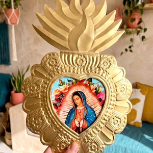 Könnte beinhalten: Ein goldfarbenes Wandornament aus Metall in Form eines heiligen Herzens mit einer Flamme an der Spitze. Die Mitte zeigt ein farbenfrohes Bild der Jungfrau von Guadalupe mit blauem Mantel und gefalteten Händen, umgeben von einem floralen Rand.