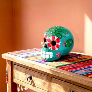 May include: A turquoise ceramic skull sculpture featuring hand-painted floral designs in red, white, and green, with a prominent green cactus motif on the forehead and sides, resting on a colorful striped textile runner atop a wooden table.