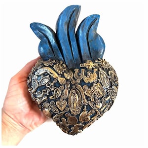 May include: A blue and gold heart-shaped religious folk art piece. The heart is adorned with various religious symbols and figures. The top of the heart features blue flame-like accents. The piece is held in a hand.