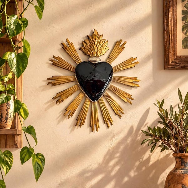 Puede incluir: Un adorno de pared con forma de corazón sagrado que presenta un corazón negro brillante en el centro, rodeado por un marco dorado radiante con rayos puntiagudos y un detalle de llama dorada estilizada en la parte superior.