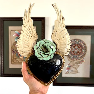 Peut inclure: Décoration murale en forme de cœur noir avec bordure dorée, ailes crème et fleur turquoise. Les ailes sont derrière le cœur, et la fleur est au-dessus. L'œuvre est tenue devant des œuvres encadrées.