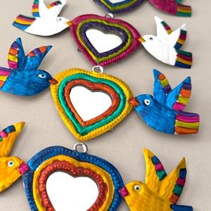 May include: Colorful metal heart-shaped ornaments with mirrors, each featuring a bird on either side. The birds are painted in vibrant hues of blue, yellow, white, and pink with rainbow-colored wings. The hearts have a layered design with a heart-shaped mirror in the center.