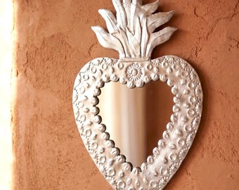 May include: A heart-shaped wall mirror with a metallic silver frame featuring embossed circular and floral patterns. The top of the frame is adorned with a stylized flame or leaf-like crest. The mirror surface is smooth and reflective.