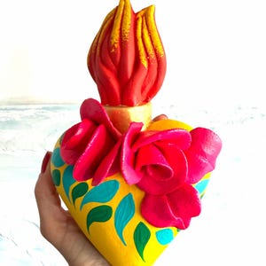 May include: A heart-shaped sculpture painted bright yellow with teal and green leaf patterns. The top features a sculpted red and yellow flame and a cluster of three-dimensional pink roses attached to the front of the heart surface.