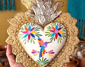 Könnte beinhalten: Ein herzförmiges Wandornament mit einem metallisch goldenen, gewellten Rand und einem weißen Mittelfeld, das mit einem bunten Kolibri und zwei Blumenmotiven bemalt ist. Die Oberseite ist mit einem strukturierten, flammenartigen Gold-Silber-Kamm verziert.