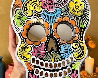 Könnte beinhalten: Ein schädelförmiger Wandspiegel mit schwarzem Hintergrund, verziert mit lebendigen, mehrfarbigen Blumen- und Schnörkelmustern in Pink, Gelb, Orange, Blau und Limettengrün. Die Augenhöhlen und die Nasenöffnung sind als reflektierende Spiegelflächen gestaltet.