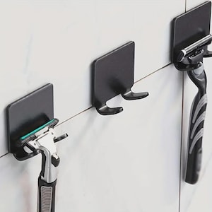 May include: Three black razor holders mounted on a white tiled wall. Two holders have razors attached, one with a green blade. The holders are square with a curved hook design for razor storage.