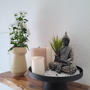 May include: A decorative shelf arrangement featuring a seated Buddha statue, candles, and a vase of white flowers. The Buddha statue is black and gold, and the candles are various shades of white and beige. The arrangement sits on a black pedestal tray with white pebbles.
