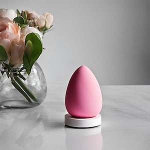 May include: A pink, egg-shaped beauty blender sponge sits on a white and pink base. A clear glass vase with pink and white roses and green leaves is in the background.