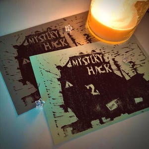 May include: Two printed cards with the text "MYSTERY HACK" and a house graphic. One card is gray, the other is mint green. A lit candle in a glass jar sits in the background, casting a warm glow. Small star-shaped decorations are scattered around the cards.
