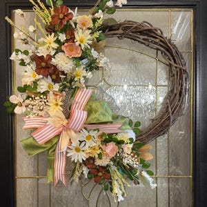 Puede incluir: Una corona circular de sarmientos adornada con un arreglo de flores artificiales en tonos óxido, crema, melocotón y amarillo, acentuada con follaje verde. Se incluye un lazo decorativo con rayas color melocotón y blanco y cinta verde.