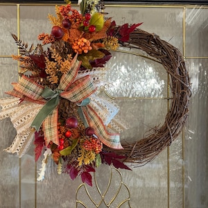 May include: A circular grapevine wreath decorated with autumn-themed elements including dark red and orange berries, pinecones, and fall-colored leaves. A multi-layered bow made of plaid fabric, lace ribbon, and a solid green velvet ribbon is attached.
