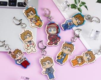 May include: A collection of seven acrylic keychains featuring cartoon-style characters with brown hair and glasses, each attached to a silver-toned crescent moon clasp. The characters are depicted in various outfits, including a red space suit and casual wear.