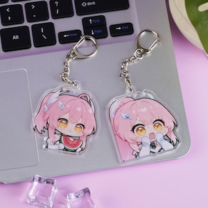 Puede incluir: Dos llaveros de estilo anime con personajes de pelo rosa. Un llavero presenta un personaje sosteniendo una rodaja de sandía. Ambos tienen llaveros y cadenas plateadas. El fondo incluye un portátil y una superficie rosa.