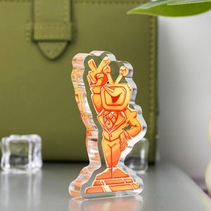 May include: A clear acrylic figurine of a cartoon character with a television for a head, wearing an orange suit and giving a peace sign. The figurine is standing on a rectangular base. The background includes a green leather wallet and a white vase.