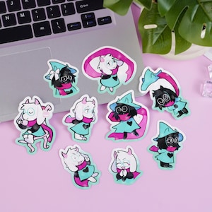 May include: A collection of colorful character stickers featuring a cartoon style. The stickers depict various characters with black, white, pink, and teal color schemes. The stickers are arranged on a pink surface near a laptop.