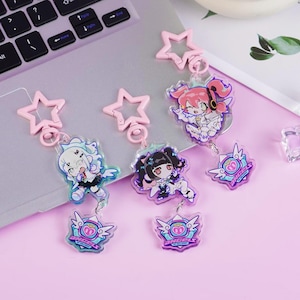Pode incluir: Três chaveiros de personagens em estilo anime com fechos em forma de estrela rosa. Cada chaveiro apresenta um design de personagem diferente com cores vibrantes e um emblema em forma de coração. O fundo é um laptop e uma superfície rosa.