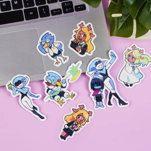 Puede incluir: Una colección de pegatinas de personajes de dibujos animados coloridos. Las pegatinas presentan varios personajes con diseños distintos y colores vibrantes, incluyendo azul, amarillo, rojo y blanco. Están dispuestas sobre un portátil y una superficie rosa.