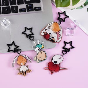 Puede incluir: Colección de cinco llaveros de personajes con cierres negros en forma de estrella. Los llaveros presentan personajes de dibujos animados coloridos con varios diseños. El fondo es una superficie rosa claro con un portátil y elementos decorativos.