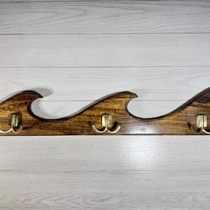 May include: A horizontal wall-mounted coat rack crafted from dark stained wood, featuring a decorative wave-shaped top edge. The rack includes three double-pronged brass-toned metal hooks evenly spaced along the front surface for hanging items.