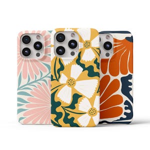 May include: Three phone cases with floral designs. One case has a pink and white floral pattern on a light blue background. Another has white flowers on a yellow background with green leaves. The third case has orange flowers on a navy background.
