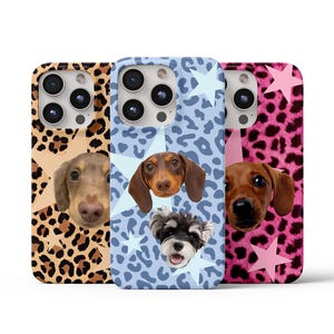 Può includere: Tre custodie per telefono con ritratti di cani. Una ha una stampa leopardata marrone e beige con un bassotto. Un'altra ha uno sfondo blu con stelle e un bassotto e uno schnauzer. La terza ha una stampa leopardata rosa con un bassotto.