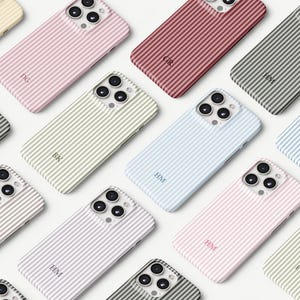 Puede incluir: Una colección de fundas para teléfono a rayas en varios colores pastel, incluyendo rosa, azul y verde. Cada funda presenta un monograma personalizado en un color contrastante. Las fundas están dispuestas en un patrón de cuadrícula, mostrando las diferentes opciones de color.