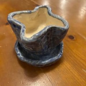 May include: A small, handmade ceramic planter with a star-shaped opening and a matching saucer. The planter is glazed in shades of blue, with a lighter interior. The piece sits on a wooden surface.