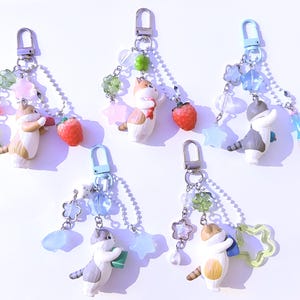 May include: Five keychains featuring small cat figurines in various poses, each with a different accessory. The keychains have silver-tone hardware and are adorned with beads, flowers, and charms in pastel colors, including pink, green, and blue.