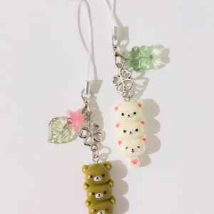 May include: Two decorative phone charms. One features three stacked olive-green bear figures, the other has three stacked white bear figures. Both charms have a silver-colored clasp and a white cord. The charms also have decorative elements like a leaf, a clover, and a pink bow.