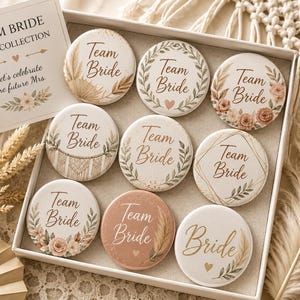 Può includere: Un set di nove spille rotonde in una scatola bianca, ognuna con la scritta Team Bride in vari disegni floreali, a foglie e geometrici. È inclusa una carta quadrata con il testo Team Bride Boho Collection Let's celebrate the future Mrs.
