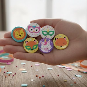 Könnte beinhalten: Eine Sammlung bunter runder Knöpfe mit Tiermotiven, darunter ein Löwe, eine Eule, ein Fuchs und ein Einhorn. Die Knöpfe werden in einer Hand gehalten, mit Konfetti und Luftschlangen auf einer Holzoberfläche. Jeder Knopf hat einen Durchmesser von etwa 3,8 cm.