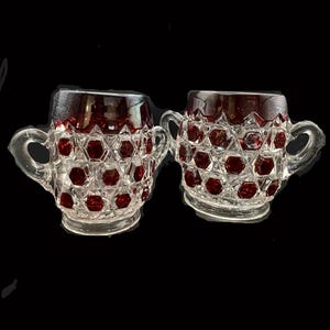 May include: Two clear glass mugs with ruby red interiors and a honeycomb pattern. Each mug has a handle and a decorative design. The mugs are likely vintage or antique and are suitable for serving beverages.
